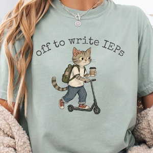 Könnte beinhalten: Hellgrünes T-Shirt mit dem Text "off to write IEPs" und einer Cartoon-Katze auf einem Roller. Die Katze hat einen Rucksack und einen Kaffeebecher. Die Katze trägt eine Schleife und rote Turnschuhe.