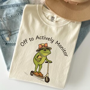Könnte beinhalten: Cremefarbenes T-Shirt mit einem Cartoon-Frosch auf einem Roller, der einen Hut und Perlen trägt und ein Buch hält. Der Text "Off to Actively Monitor" ist um den Frosch herum gedruckt. Das Shirt wird mit einer Jeans und einem beigen Hut kombiniert.