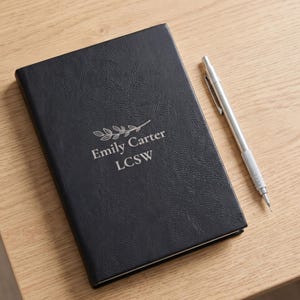 May include: A black notebook with a textured cover featuring a silver engraved branch graphic above the text Emily Carter LCSW, accompanied by a silver mechanical pencil with a clip and a pointed tip resting beside the notebook.