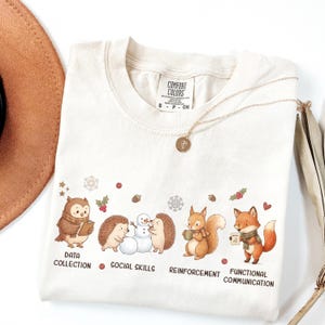 May include: A cream-colored t-shirt featuring a graphic of an owl, two hedgehogs building a snowman, a squirrel, and a fox. Text below the animals reads DATA COLLECTION, SOCIAL SKILLS, REINFORCEMENT, and FUNCTIONAL COMMUNICATION.