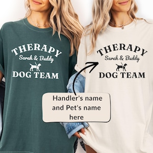 Könnte beinhalten: Zwei T-Shirts, eines dunkelgrün und eines cremefarben, jeweils mit dem Text "THERAPY Sarah & Buddy DOG TEAM" in Weiß. Eine Grafik eines Hundes befindet sich zwischen "Buddy" und "Dog Team". Der untere Teil des Bildes hat ein Textfeld mit der Aufschrift "Handler's name and Pet's name here".