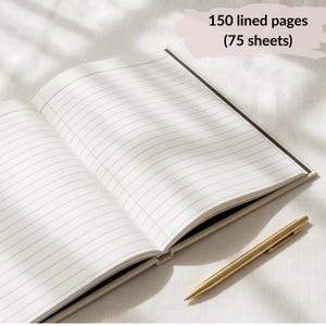 May include: An open notebook with white lined pages and a gold-toned metal pen. The notebook features 150 lined pages (75 sheets) as indicated by the text overlay. The pages are white with thin black horizontal lines, and the pen has a slim, metallic finish.