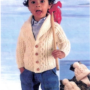 May include: A young child wearing a white cable knit cardigan with a shawl collar and button closure. The child is also wearing blue jeans and a brown bandana with a white pattern. The child is holding a wooden stick with a red fabric decoration attached to it. There are two white knitted hats in the background.