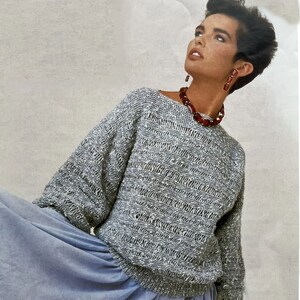May include: A grey and white knitted jumper with a loose fit and long sleeves. The jumper has a ribbed texture and a round neckline. The model is wearing a blue skirt and a red beaded necklace.