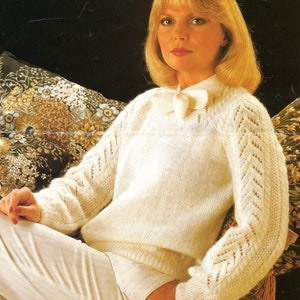 Könnte beinhalten: Ein weißer Strickpullover mit zartem Spitzenmuster und einer Schleife am Ausschnitt. Der Pullover wird über einer weißen Hose getragen.