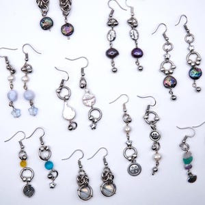 Puede incluir: Una colección de varios pendientes colgantes con ganchos, cadenas y anillos de metal plateado. Los diseños incorporan diversos elementos, incluyendo cuentas de vidrio iridiscente, pequeñas perlas blancas, botones de metal, dijes en forma de estrella y cuentas de piedras preciosas.