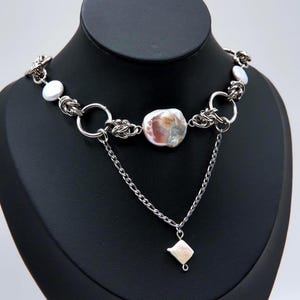 Puede incluir: Un collar de cadena plateada con múltiples eslabones circulares intercalados con perlas blancas iridiscentes. Una perla central irregular más grande está flanqueada por anillos metálicos, con una cadena colgante delicada y un pequeño dije de perla cuadrada.