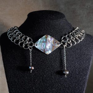 Puede incluir: Un collar con un colgante central en forma de diamante de concha de abulón iridiscente, flanqueado por eslabones de cadena plateados. Dos delicadas cadenas cuelgan del colgante, cada una terminada en una pequeña cuenta redonda oscura.