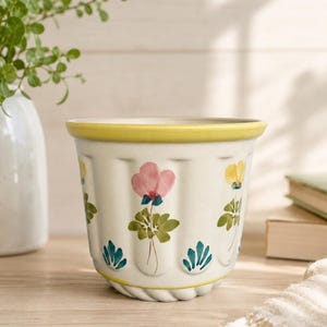 May include: A cream-coloured ceramic planter with a fluted, ribbed exterior and a yellow rim. The surface features hand-painted floral motifs in pink, yellow, and green, interspersed with small blue leaf-like accents around the base.