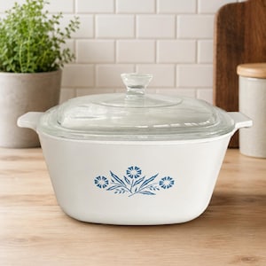 Puede incluir: Una fuente de horno cuadrada de color blanco con esquinas redondeadas y asas laterales integradas. El exterior presenta un estampado floral azul en la parte frontal. Incluye una tapa de cristal transparente con un pomo central para levantarla.
