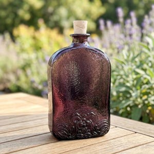 Peut inclure: Une bouteille en verre violet foncé avec une surface texturée et un bouchon en liège. La bouteille présente une forme rectangulaire arrondie avec un motif en relief décoratif orné gravé le long du bord inférieur de la face avant.