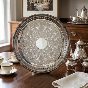 May include: A large, round silver serving tray featuring an intricate, embossed floral and scrollwork pattern surrounding a plain, circular centre. The tray has a raised, rope-style border with a decorative pierced edge design.