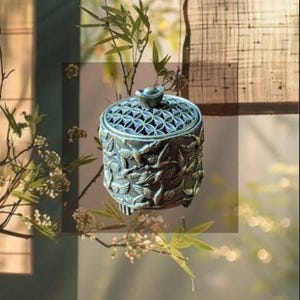 May include: A cylindrical metal incense burner featuring an intricate relief pattern of flying cranes on the exterior, a geometric lattice lid with a small knob handle, and three short feet, presented in a dark, weathered metallic finish.