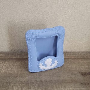 May include: A light blue square picture frame featuring a matte finish and a raised white cameo relief at the bottom center. The frame is decorated with a beaded border and small bow accents at the top corners, creating a textured appearance.