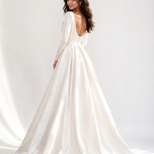May include: A floor-length ivory gown with long sleeves and a deep scoop back. The dress features a fitted bodice that transitions into a full, pleated skirt with a sweeping train, crafted from a smooth, slightly lustrous fabric.