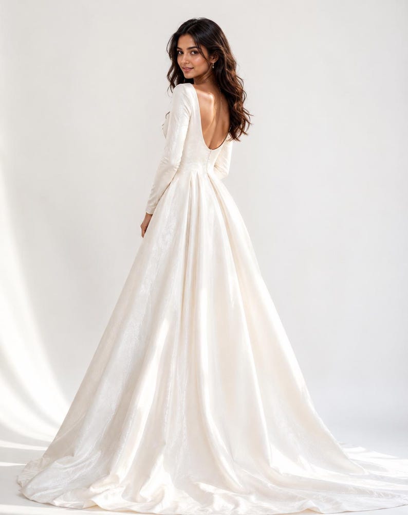 May include: A floor-length ivory gown with long sleeves and a deep scoop back. The dress features a fitted bodice that transitions into a full, pleated skirt with a sweeping train, crafted from a smooth, slightly lustrous fabric.