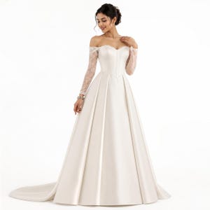 May include: An off-the-shoulder floor-length gown in ivory satin featuring long sheer lace sleeves, a sweetheart neckline, a fitted bodice with vertical seam detailing, and a full pleated skirt that extends into a long train on the floor.