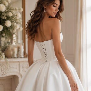 Op de afbeelding: Een strapless witte baljurk met een gestructureerd korset-lijfje met een vetersluiting aan de achterkant. De jurk heeft een volle, volumineuze rok van gladde stof die tot op de grond valt, wat zorgt voor een klassiek silhouet met strakke lijnen.