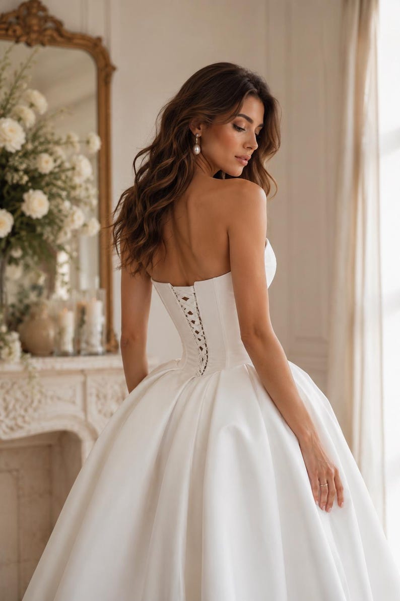 Op de afbeelding: Een strapless witte baljurk met een gestructureerd korset-lijfje met een vetersluiting aan de achterkant. De jurk heeft een volle, volumineuze rok van gladde stof die tot op de grond valt, wat zorgt voor een klassiek silhouet met strakke lijnen.