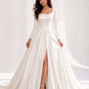 May include: A floor-length white gown with a square neckline and long sleeves. The dress features a fitted bodice with vertical seams and a full, flowing skirt that includes a high front slit, revealing the leg. The fabric has a subtle sheen.