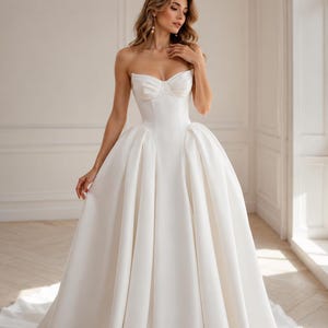 Op de afbeelding: Een vloerlange witte strapless trouwjurk met een gestructureerd sweetheart-lijfje met zichtbare naaddetails en een volumineuze, geplooide rok die vanaf de taille uitloopt in een volle, slepende rok gemaakt van gladde, effen stof.