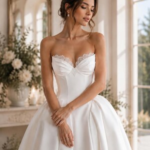 Op de afbeelding: Een strapless witte japon met een gestructureerd lijfje met een hartvormige halslijn en een verfijnde kanten rand langs de boord. De jurk heeft een getailleerde taille die overgaat in een volle, volumineuze rok met soepele plooien.