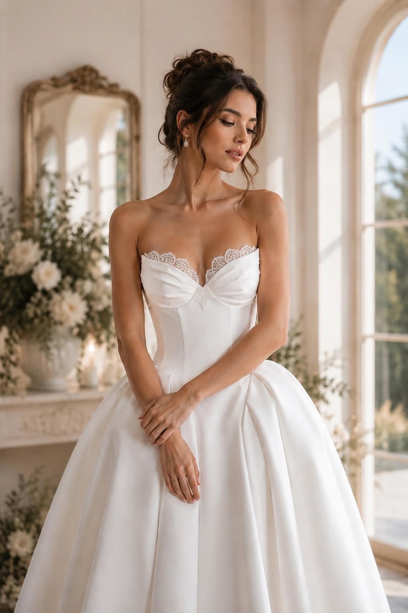 Op de afbeelding: Een strapless witte japon met een gestructureerd lijfje met een hartvormige halslijn en een verfijnde kanten rand langs de boord. De jurk heeft een getailleerde taille die overgaat in een volle, volumineuze rok met soepele plooien.