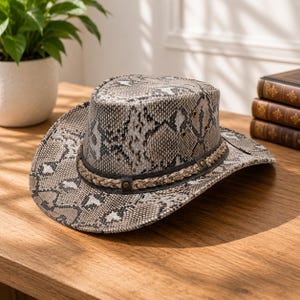 Può includere: Un cappello da cowboy con motivo a pelle di serpente grigio e bianco. Il cappello presenta una fascia intrecciata marrone scuro attorno alla base della corona, arricchita da piccoli borchie metalliche e due occhielli di ventilazione circolari.