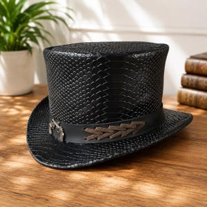 May include: A black top hat featuring a textured snakeskin pattern across the crown and brim. A smooth black leather band encircles the base, decorated with a silver-toned metal pistol emblem and a brown suede lace-up detail on the side.