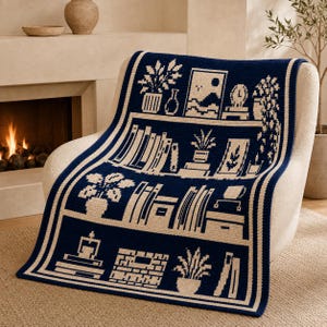 May include: A dark blue and cream crochet blanket featuring a repeating bookshelf pattern. The design includes rows of books, potted plants, framed art, and decorative objects. The blanket has a thick border and a textured, woven appearance.