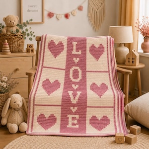 May include: A cream and pink crocheted blanket draped over a wooden chair. The blanket features a repeating pattern of pink hearts and the word "LOVE" in pink letters. The blanket is a cozy, handmade item.