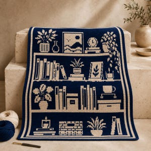 May include: A dark blue and cream crocheted blanket featuring a bookshelf pattern. The design displays three shelves filled with various items including potted plants, books, a framed picture, a clock, and a coffee mug, all rendered in a two-tone graphic style.
