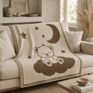 May include: A cream-colored knit blanket featuring a brown graphic design of a teddy bear sitting on a cloud. The pattern includes a crescent moon and several stars surrounding the bear. The blanket has a dark brown border along the edges.