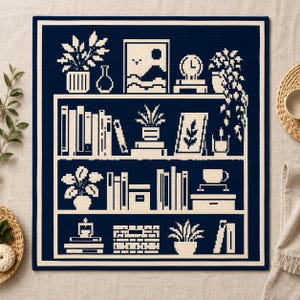 May include: A square textile wall hanging featuring a dark navy blue background with a cream-colored pixel art design of a three-tiered bookshelf. The shelves are filled with various potted plants, books, a clock, a framed picture, and a coffee cup.
