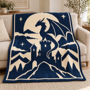 May include: A rectangular crochet blanket featuring a dark navy blue and cream color scheme. The design depicts a large dragon perched atop a castle tower against a full moon, surrounded by mountain silhouettes and small four-pointed stars.