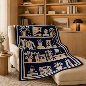 May include: A dark blue and cream knit throw blanket featuring a repeating pattern of bookshelves filled with various books, potted plants, a clock, and decorative items. The design is structured with horizontal shelves and a thick border.