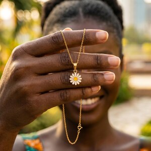 May include: A gold-colored necklace with a daisy pendant. The daisy has white petals and a yellow center. The necklace is held up in front of a person's face, with the chain and clasp visible. The background is blurred with green and yellow tones.
