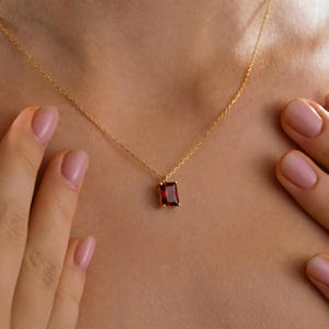 May include: A gold necklace with a rectangular, deep red gemstone pendant. The pendant is set in a gold frame and hangs from a delicate gold chain. The necklace is displayed on a person's neck.