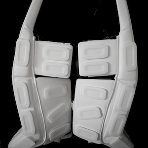 May include: Pair of white protective sports pads, possibly for ice hockey or similar sports. The pads have a series of rectangular, raised sections and long, padded straps. The background is black.