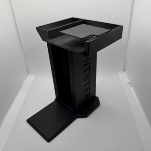 K&ouml;nnte beinhalten: Ein schwarzer, 3D-gedruckter St&auml;nder mit rechteckiger Basis, vertikaler St&uuml;tze und oberer Ablage. Das Wort "HITSTER" ist auf der St&uuml;tze gepr&auml;gt. Der St&auml;nder hat ein modernes, minimalistisches Design.