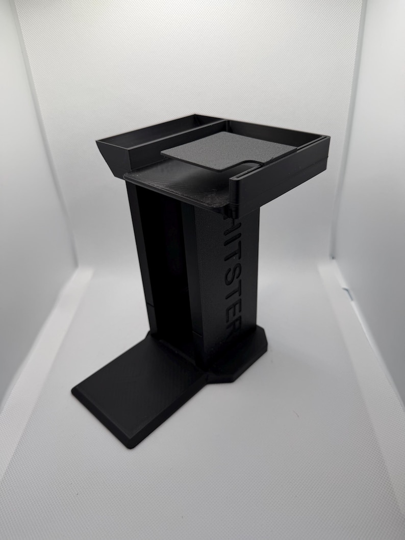 K&ouml;nnte beinhalten: Ein schwarzer, 3D-gedruckter St&auml;nder mit rechteckiger Basis, vertikaler St&uuml;tze und oberer Ablage. Das Wort "HITSTER" ist auf der St&uuml;tze gepr&auml;gt. Der St&auml;nder hat ein modernes, minimalistisches Design.