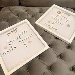 Puede incluir: Dos obras de arte con nombres familiares enmarcadas en blanco. Una dice "Family" con nombres y corazones de madera. La otra dice "Grandchildren" con nombres y un icono de casa. Ambas tienen marcos blancos y están sobre una superficie gris acolchada.
