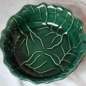 May include: A glossy, dark green ceramic bowl shaped like a cabbage leaf. The bowl features a textured surface with raised, lighter green veins that mimic the natural structure of a leaf, and a scalloped, wavy rim that follows the organic shape.