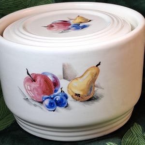 May include: A white ceramic canister with a matching lid, featuring a painted fruit motif on the front and lid. The design includes a red apple, a yellow pear, and clusters of blue grapes, all rendered in a realistic style on the smooth surface.