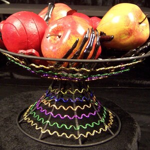 May include: A black metal pedestal bowl featuring a decorative rim and base accented with wavy gold, purple, and green tinsel. The bowl contains several red and yellow artificial apples interspersed with a black skeletal hand sculpture.