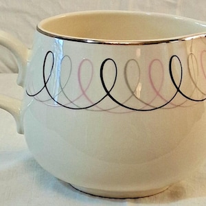May include: A cream-colored ceramic creamer featuring a curved handle and a pouring spout. The exterior is decorated with a repeating pattern of interlocking loops in black, grey, and soft pink. The rim is finished with a thin metallic silver trim.