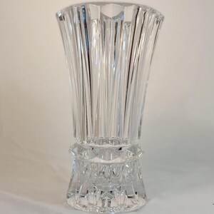 May include: A clear, cut-glass vase with a flared top and a detailed, geometric base. The vase features vertical, linear cuts that create a textured surface. The design is elegant and suitable for floral arrangements or decorative display.