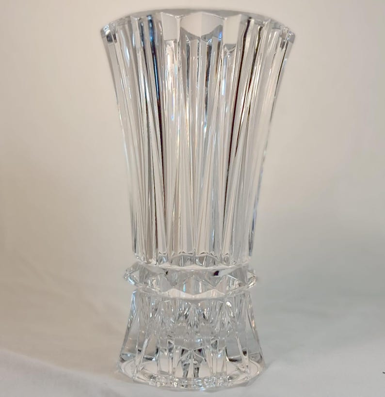 May include: A clear, cut-glass vase with a flared top and a detailed, geometric base. The vase features vertical, linear cuts that create a textured surface. The design is elegant and suitable for floral arrangements or decorative display.