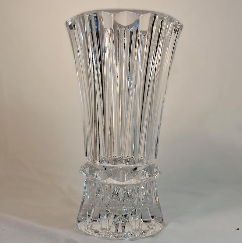 May include: A clear, faceted crystal vase with a flared top and a decorative base. The vase features vertical, linear cuts that create a textured surface. The vase is approximately 10 inches tall.