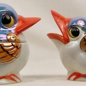 May include: A ceramic sugar bowl and creamer set shaped like birds. The sugar bowl has a blue iridescent lid, while the creamer features an open beak. Both pieces are white with orange beaks, orange feet, and gold-toned wings with black outlines.